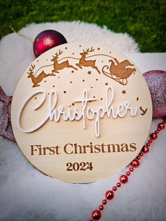 A round wooden plaque engraved with 'My First Christmas' is held by a new mother next to her baby, used as a photoshoot prop.